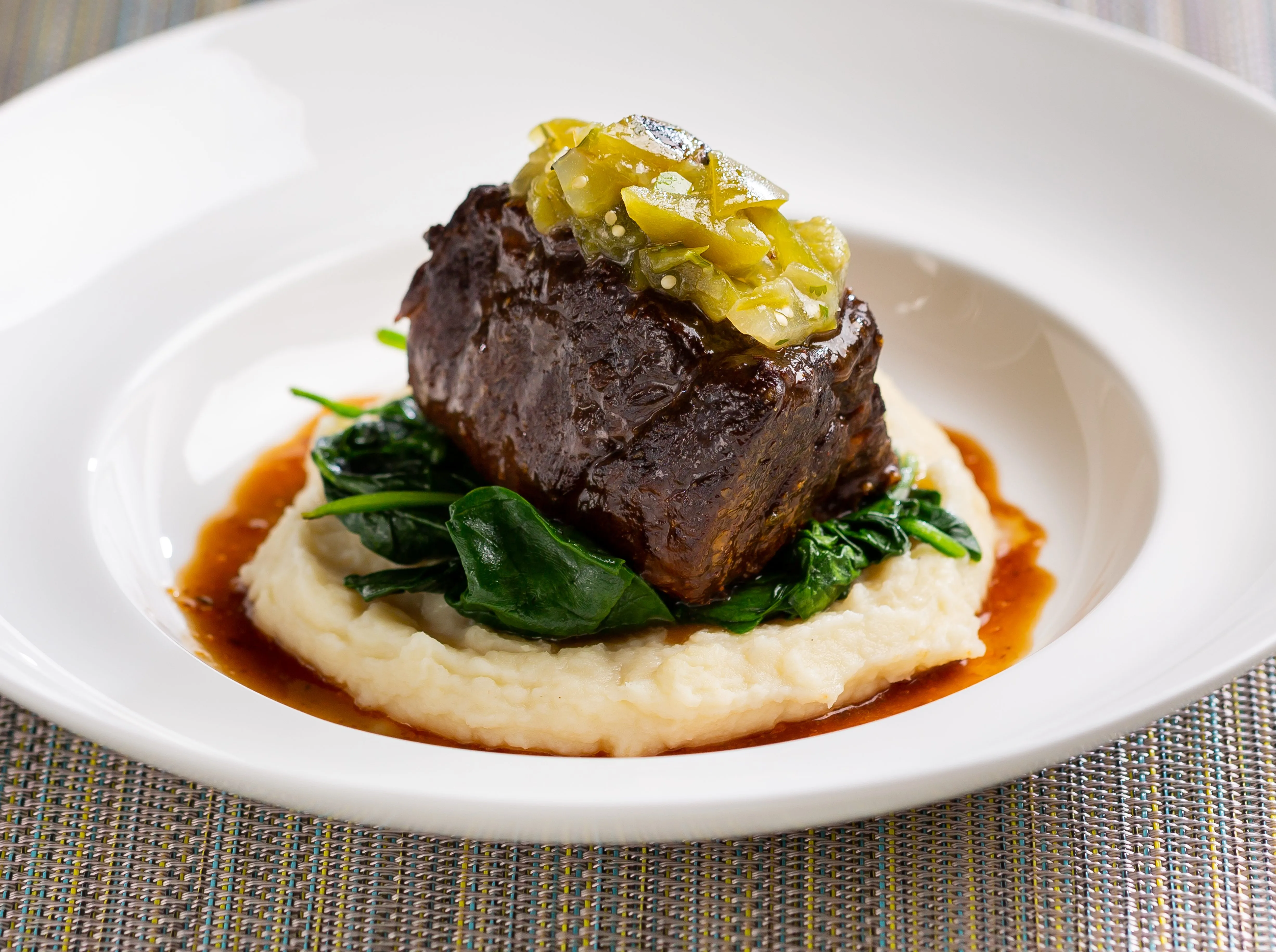 A Dutch oven filled with braised beef short ribs in a glossy, dark sauce with carrots and onions, photographed on a kitchen counter with a ladle resting nearby