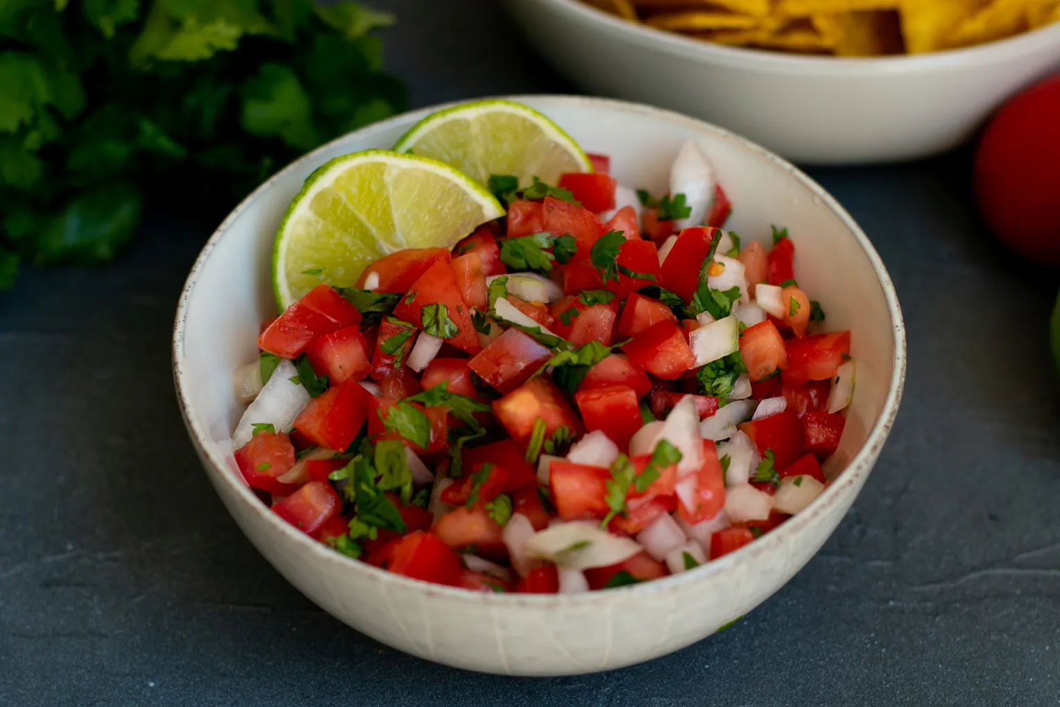 A ceramic bowl filled with fresh pico-style salsa, with visible diced tomatoes, white onion, chopped cilantro, and green chile on a kitchen counter