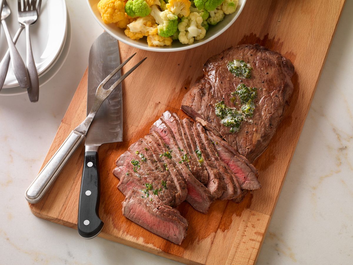 A cooked London broil on a cutting board being sliced very thin against the grain, with juices resting on the board