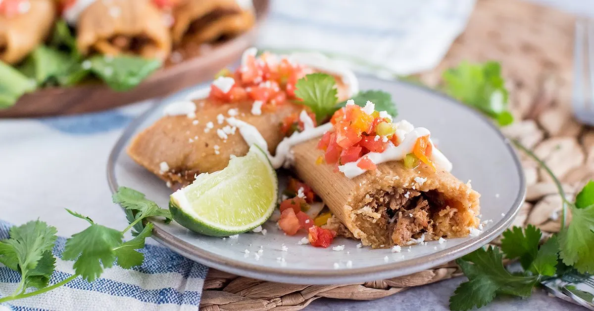 A freshly steamed tamale unwrapped on a plate with the corn husk pulled back, showing firm, fluffy masa around a shredded pork filling