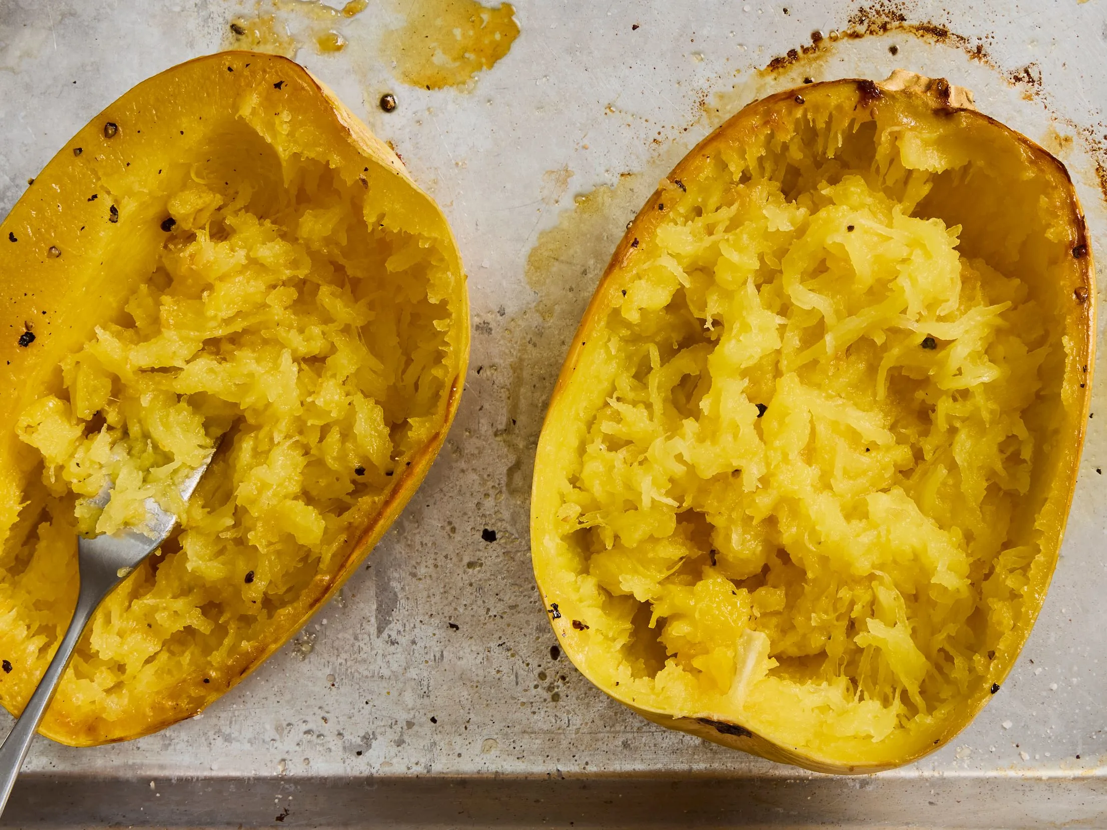 A halved roasted spaghetti squash on a sheet pan with golden edges, with strands fluffed by a fork
