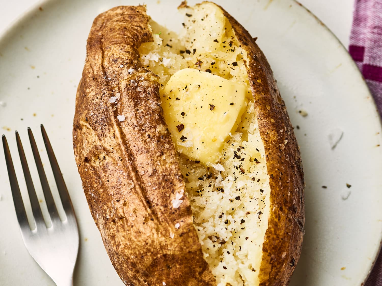 A hot microwave-baked russet potato split open on a white plate with a pat of butter melting into the fluffy center