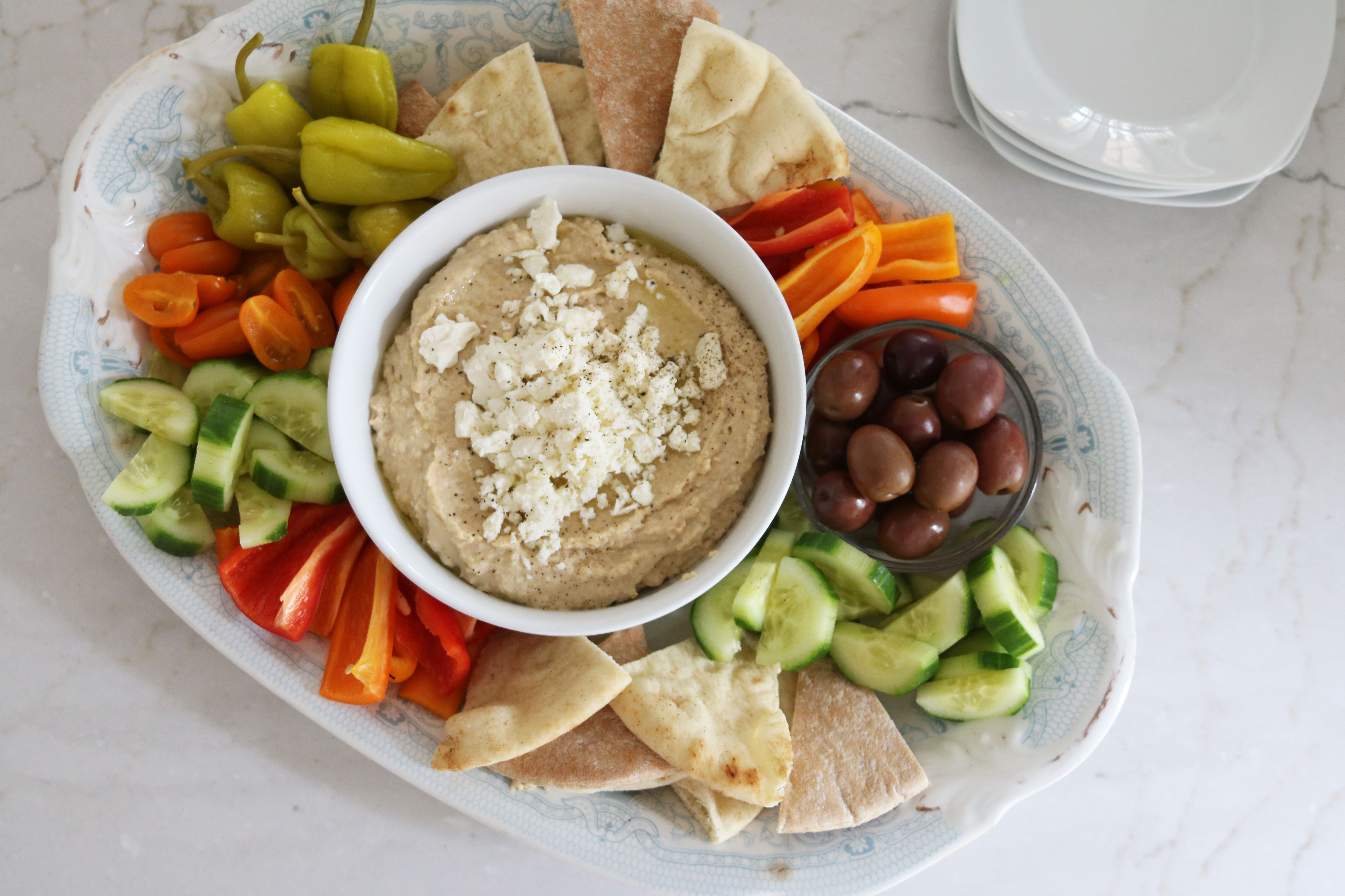 A hummus platter with a bowl of hummus surrounded by pita wedges and sliced cucumbers and carrots on a wooden board