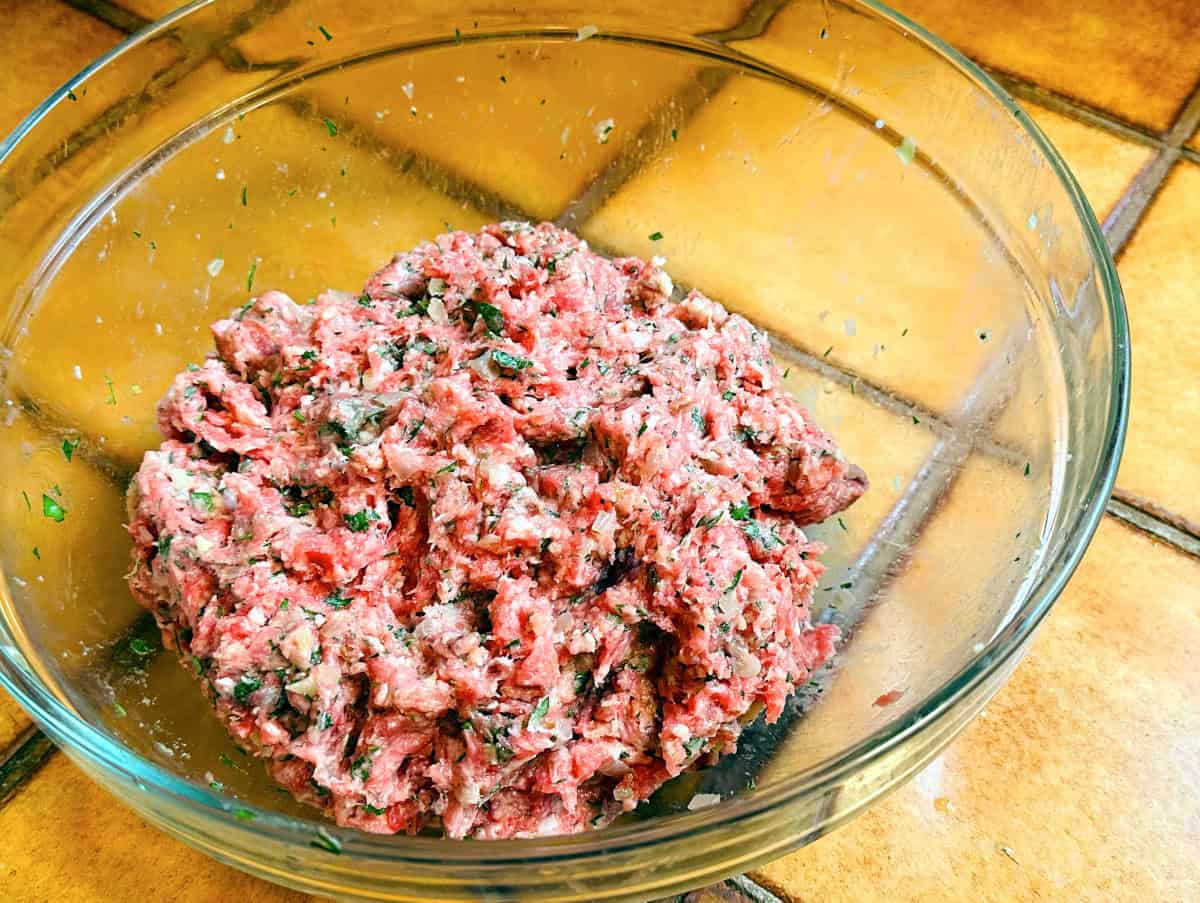 A large mixing bowl with beef and pork meatball mixture, breadcrumbs, grated onion, and parsley partially mixed with a fork