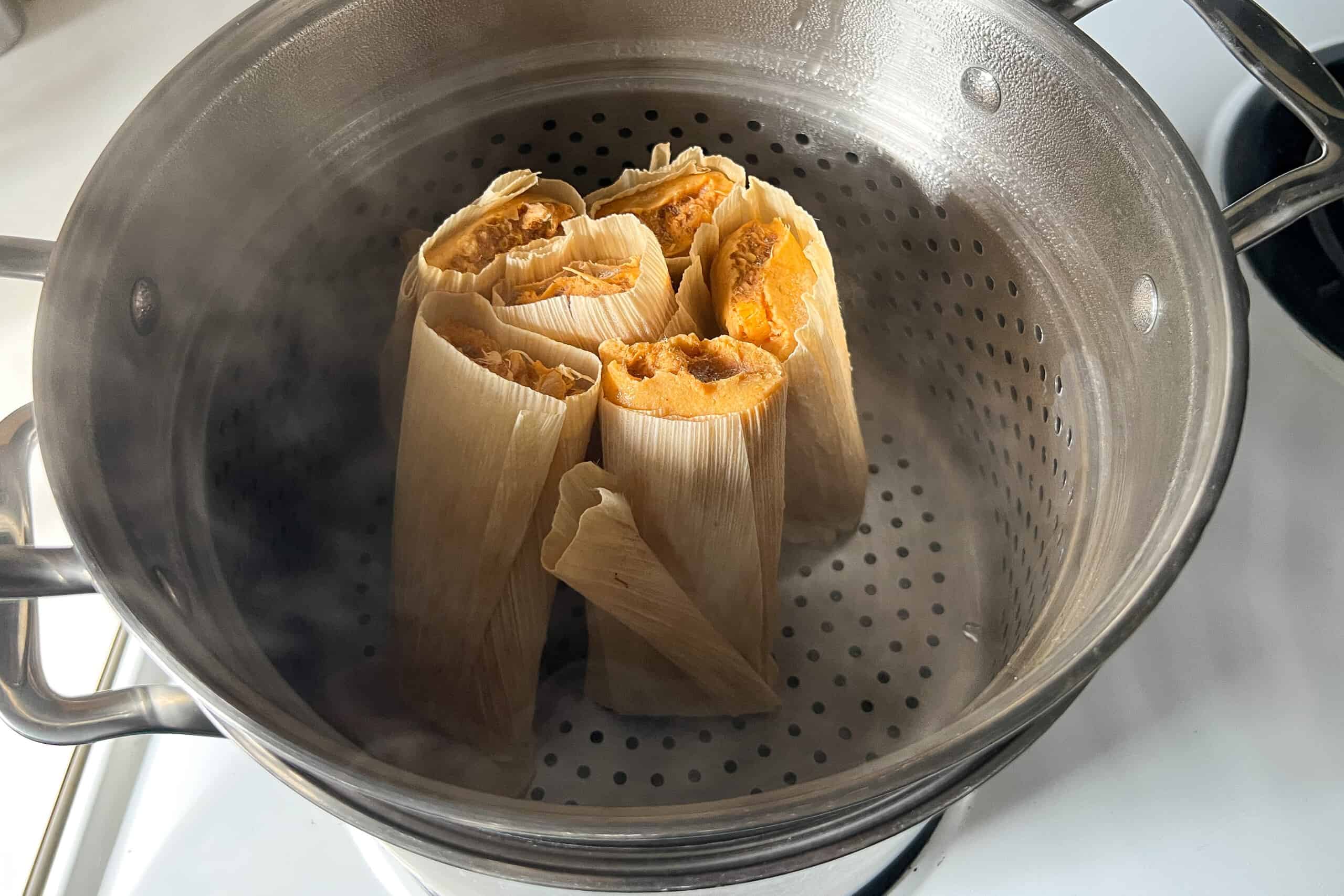 A large pot fitted with a steamer basket holding upright corn husk tamales under a lid with steam rising in a home kitchen