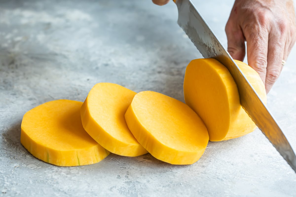 A peeled butternut squash standing upright on a cutting board while a hand uses a vegetable peeler to remove the last strip of skin