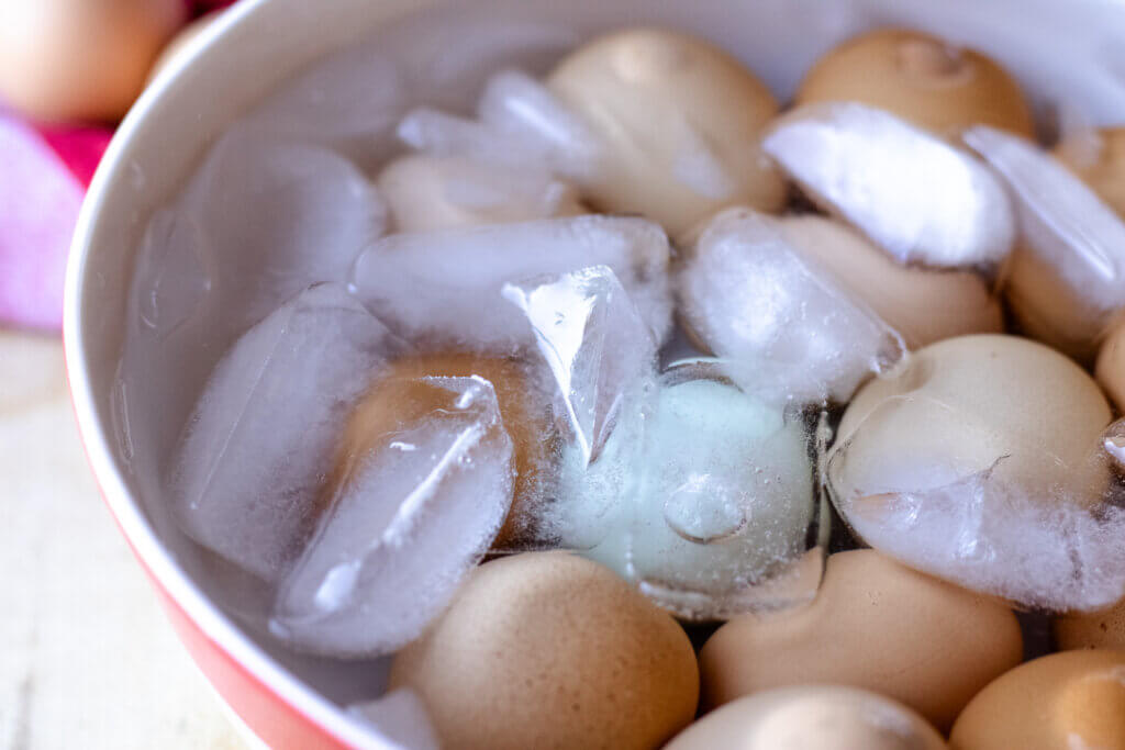 A pot of freshly cooked eggs being transferred into a bowl of ice water on a kitchen counter