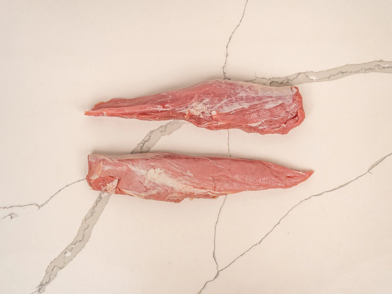 A raw pork tenderloin on a wooden cutting board with a small pile of salt and pepper and a pair of tongs beside it