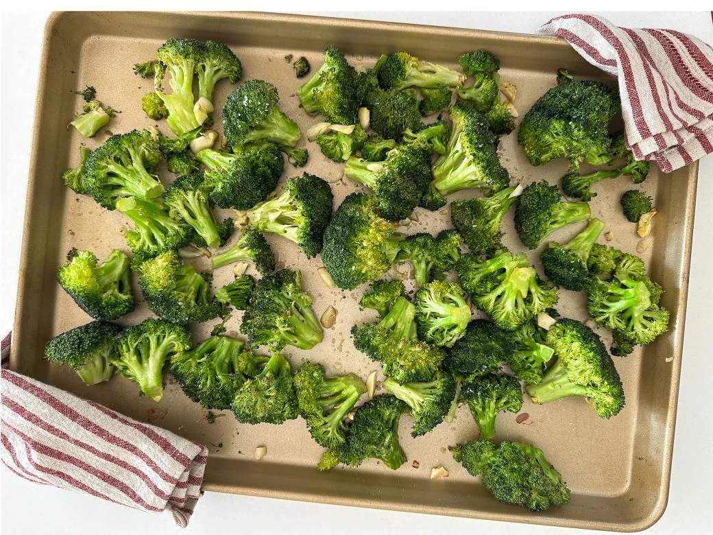 A rimmed sheet pan of roasted broccoli florets and stem pieces with browned edges and a light sheen of olive oil, fresh from the oven