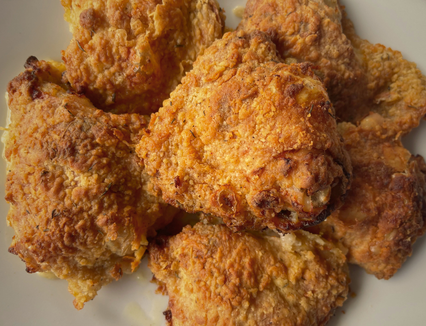 A sheet pan of golden, crispy-skinned baked chicken thighs resting in a roasting pan with pan juices, photographed in a bright home kitchen