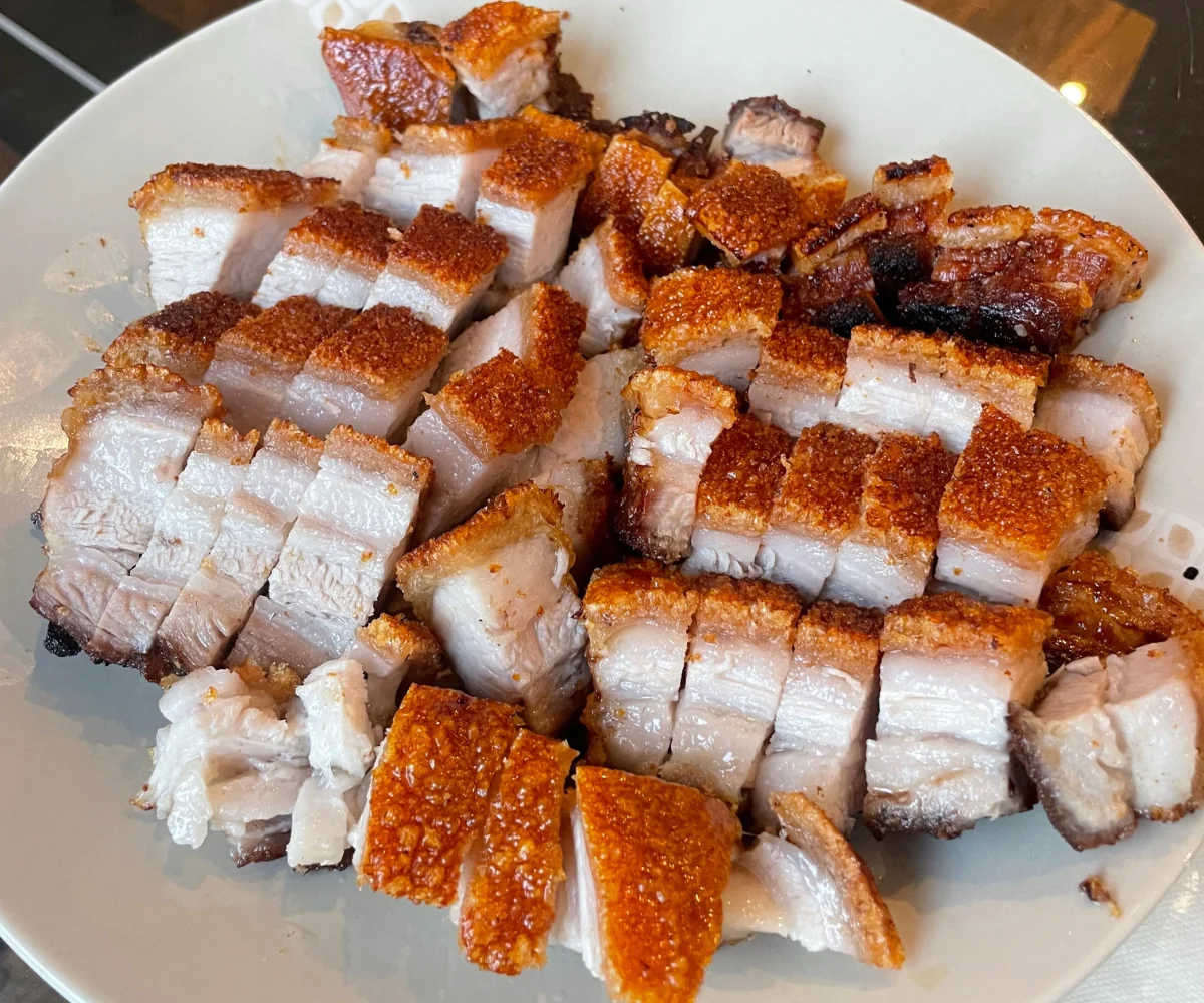 A single slab of pork belly on a rimmed baking sheet with deeply blistered, crackling skin and rendered fat pooled around it