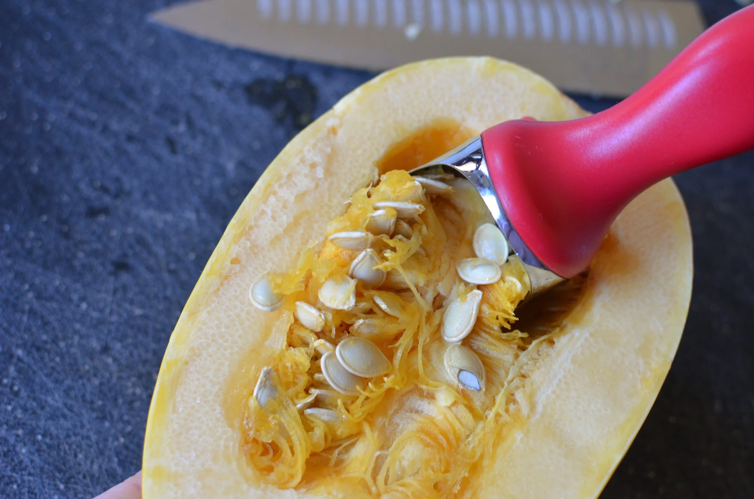 A spaghetti squash cut lengthwise on a cutting board while a hand scoops out the seeds with a metal spoon
