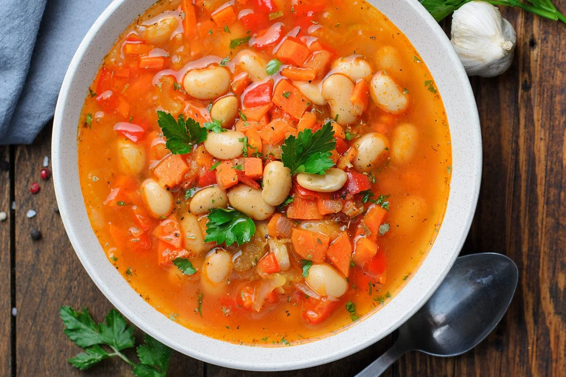A steaming bowl of classic mixed vegetable soup with carrots, celery, green beans, potatoes, and peas in a light tomato broth, photographed on a wooden table with a spoon nearby