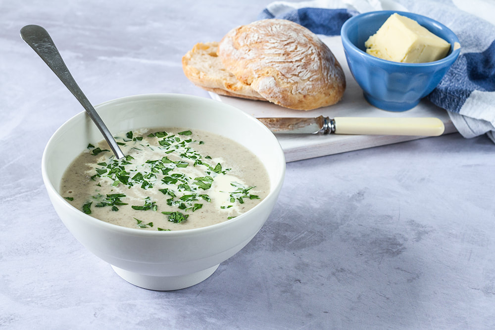 A steaming bowl of creamy mushroom soup with browned mushroom slices on top, a swirl of cream, and chopped parsley on a wooden table with a spoon beside it