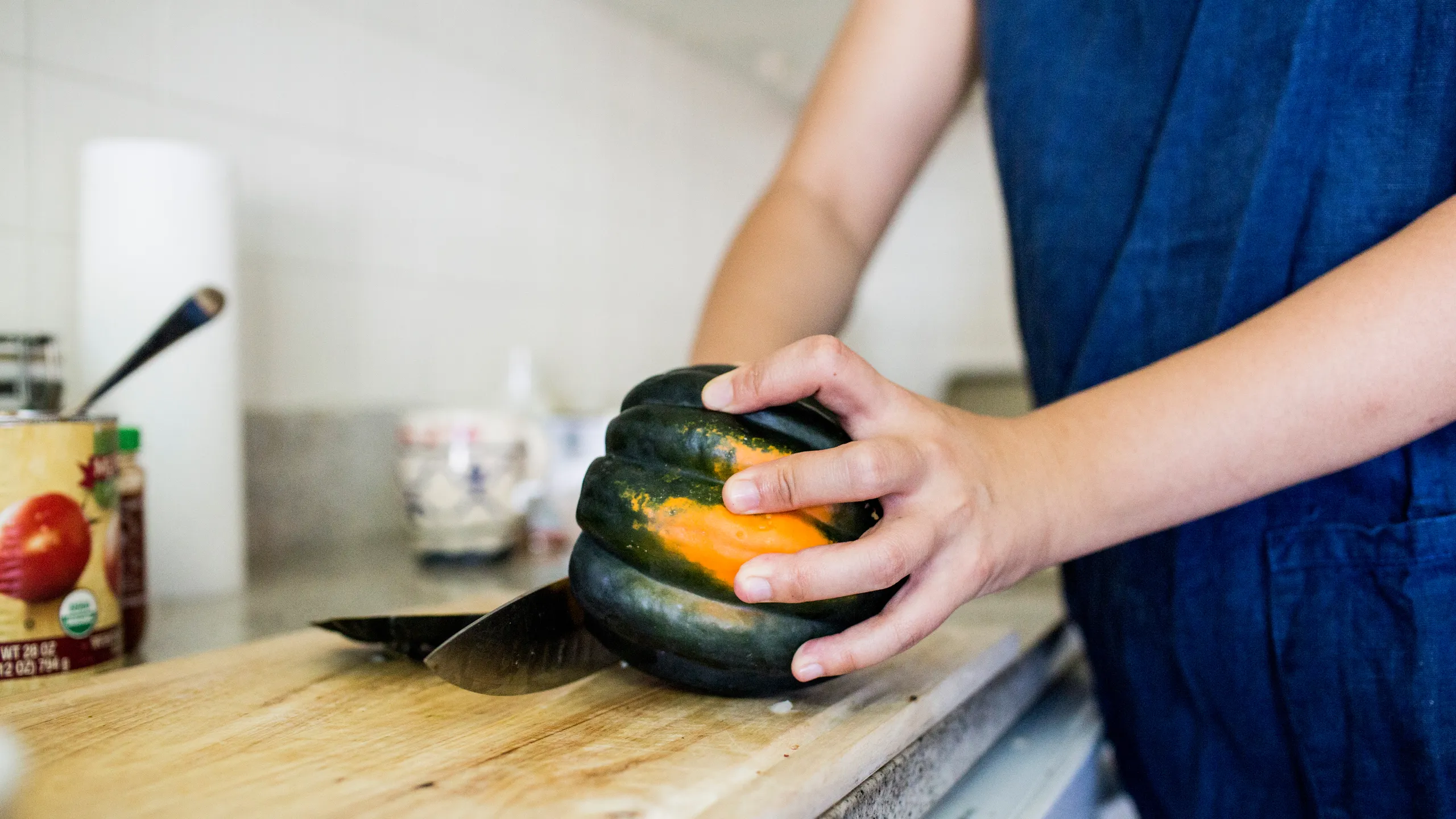 A whole acorn squash on a wooden cutting board being sliced in half with a sturdy chef's knife, hands positioned safely