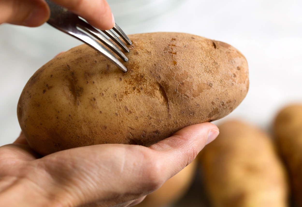 A whole russet potato being pierced several times with a fork on a cutting board
