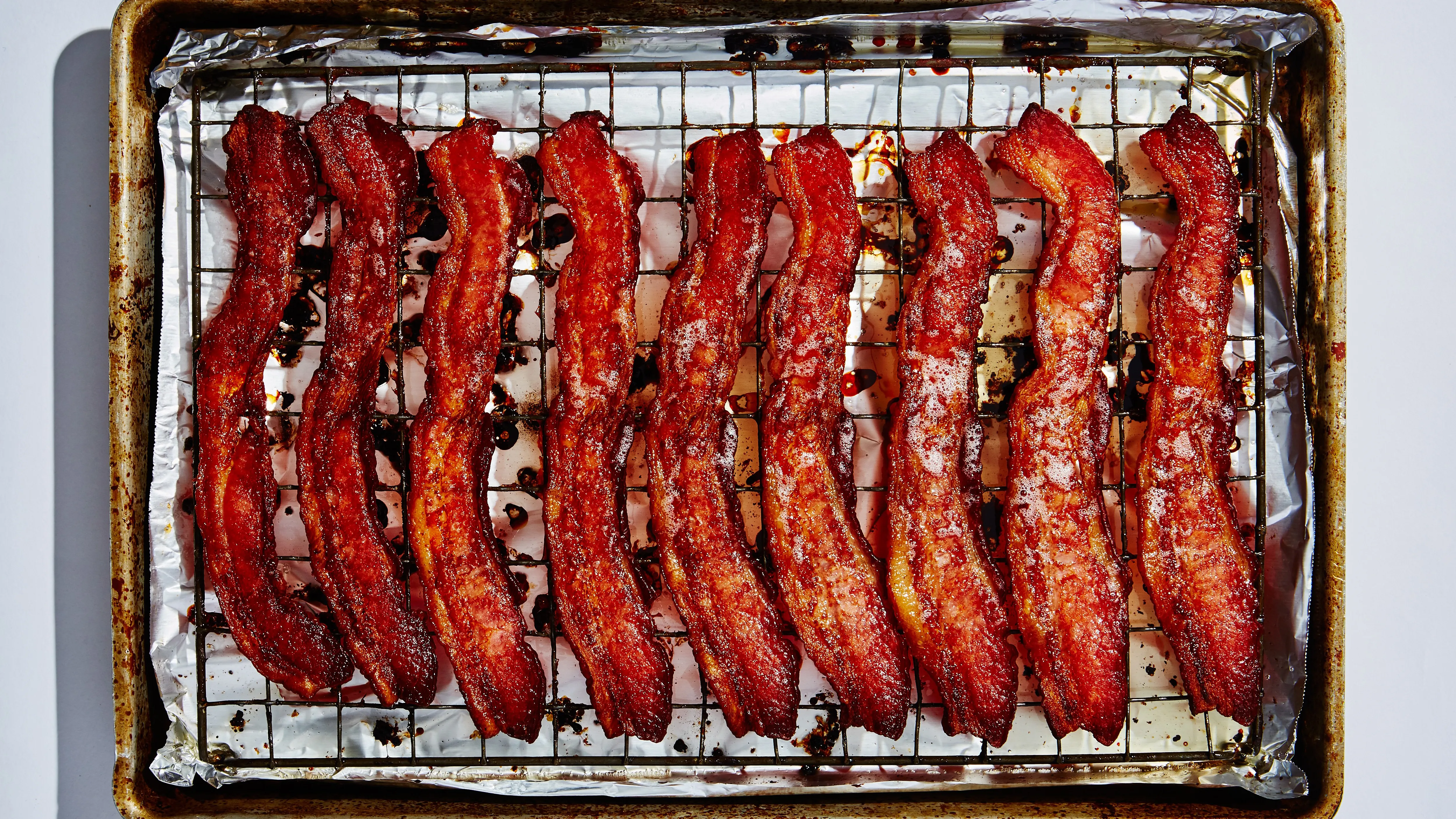 Bacon strips sizzling and browning on a foil-lined sheet pan inside an open oven, showing rendered fat pooling around the strips