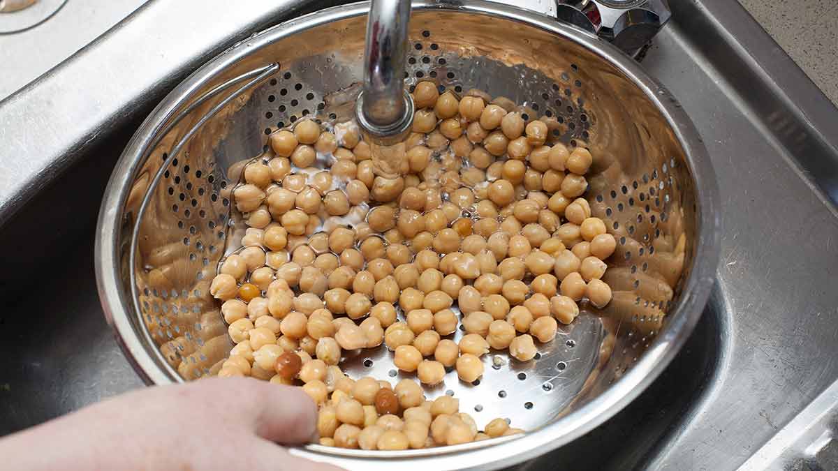 Canned chickpeas draining in a metal colander over a sink, with water droplets visible on the chickpeas