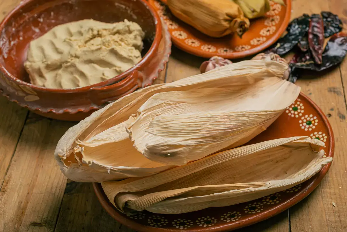 Hands folding a corn husk tamale, showing masa spread on the husk with a line of shredded pork filling before the husk is wrapped