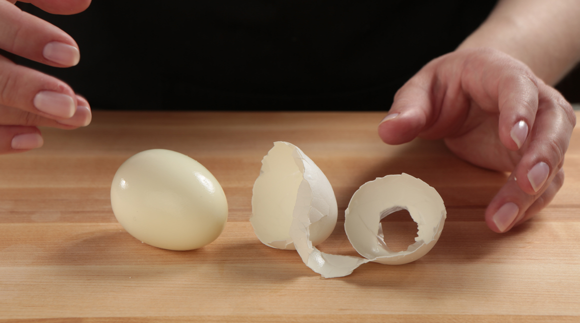 Hands peeling a hard-boiled egg over a bowl, with large shell pieces lifting cleanly from the egg white