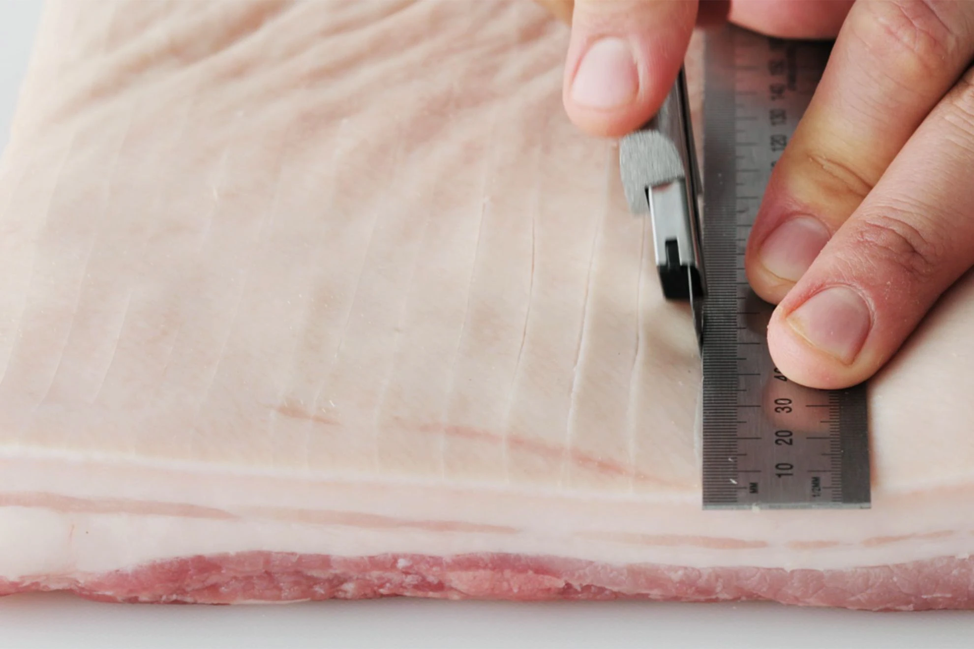 Hands scoring pork belly skin in neat parallel lines on a cutting board