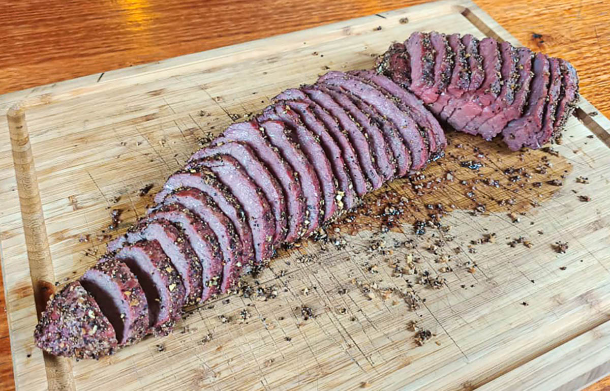 Hands slicing a rested smoked tri-tip with a chef's knife, showing the knife cutting across the grain on a cutting board