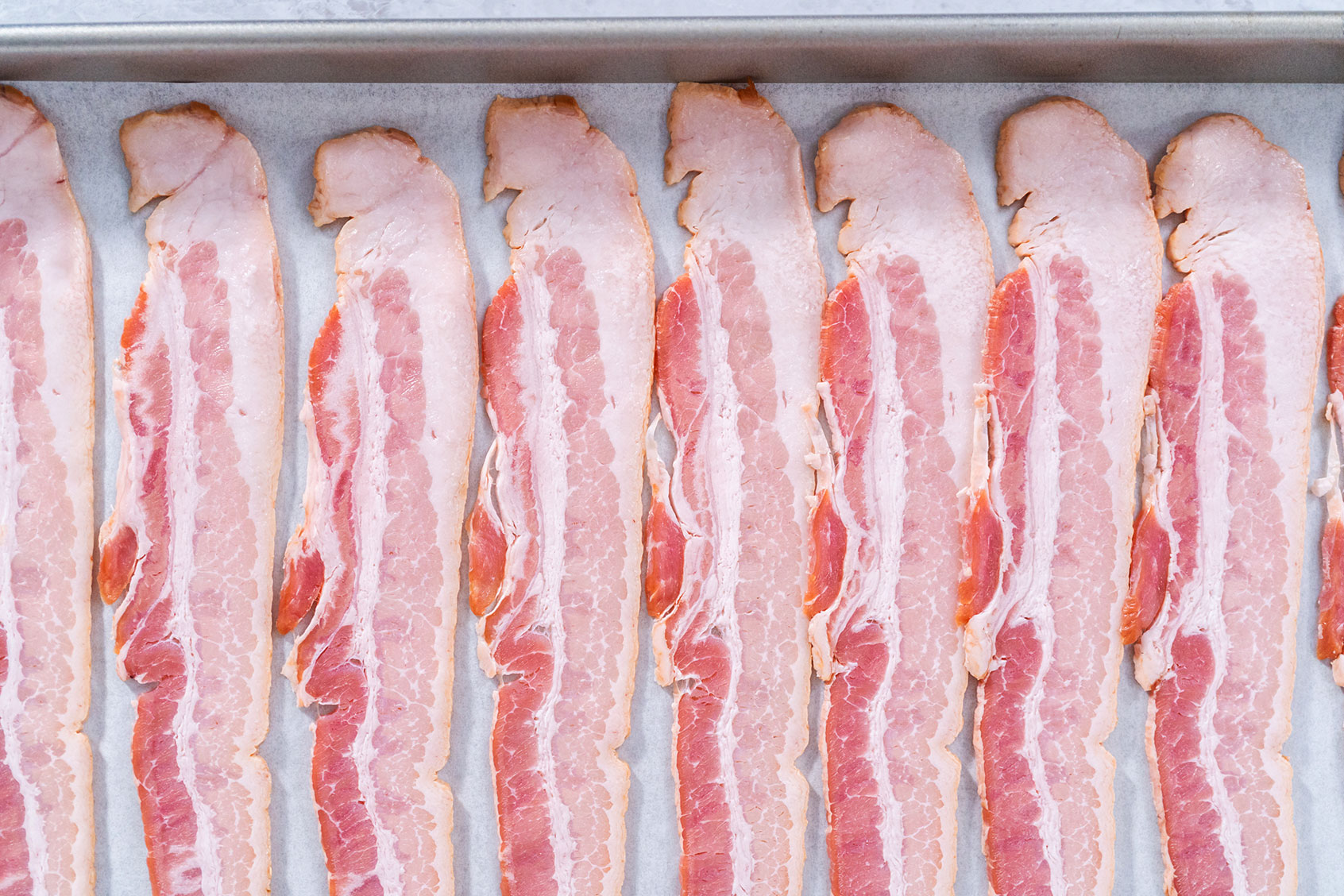 Raw bacon strips arranged in a single layer on a foil-lined rimmed sheet pan on a kitchen counter, ready to go into the oven