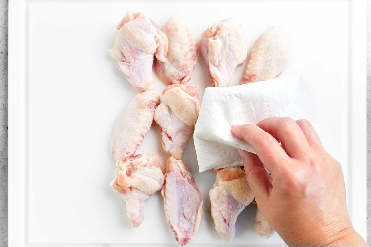 Raw chicken wings being thoroughly patted dry with paper towels on a cutting board