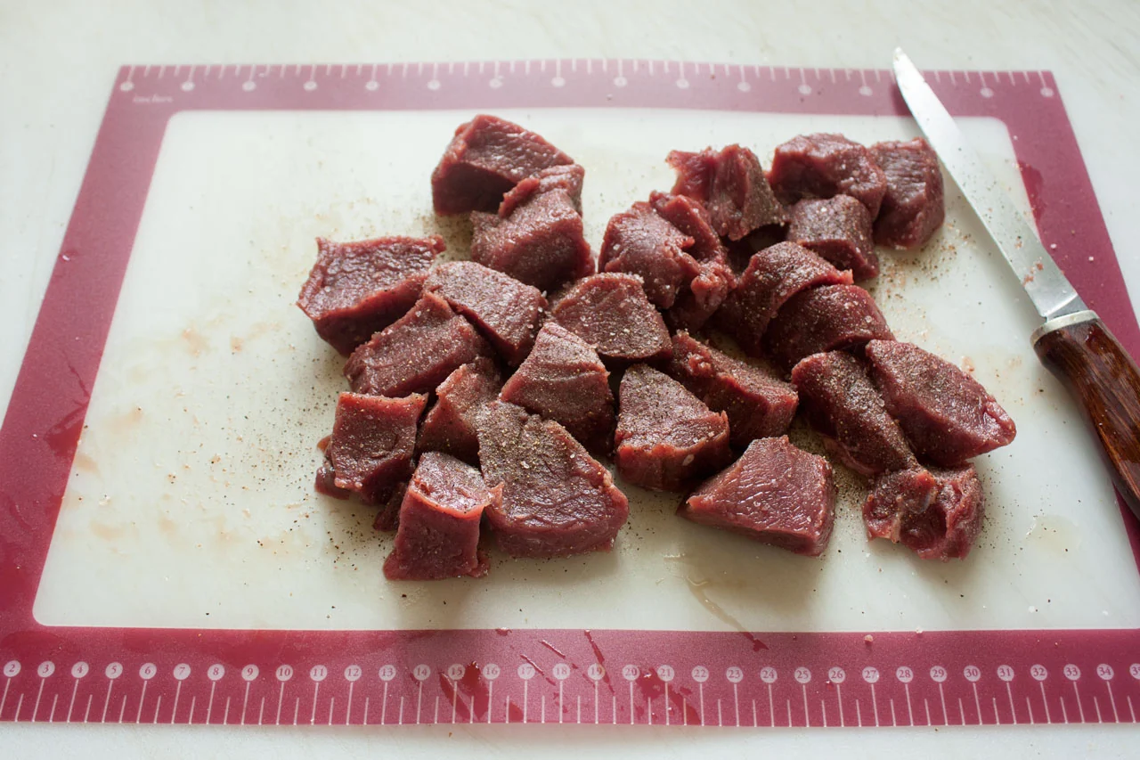 Raw venison stew meat cut into evenly sized cubes on a cutting board next to chopped onion and carrots