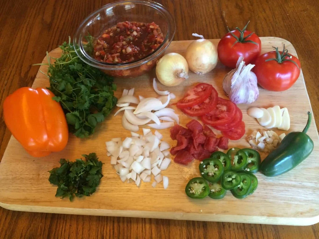 Ripe tomatoes, a white onion, cilantro, limes, and a jalapeño on a wooden cutting board
