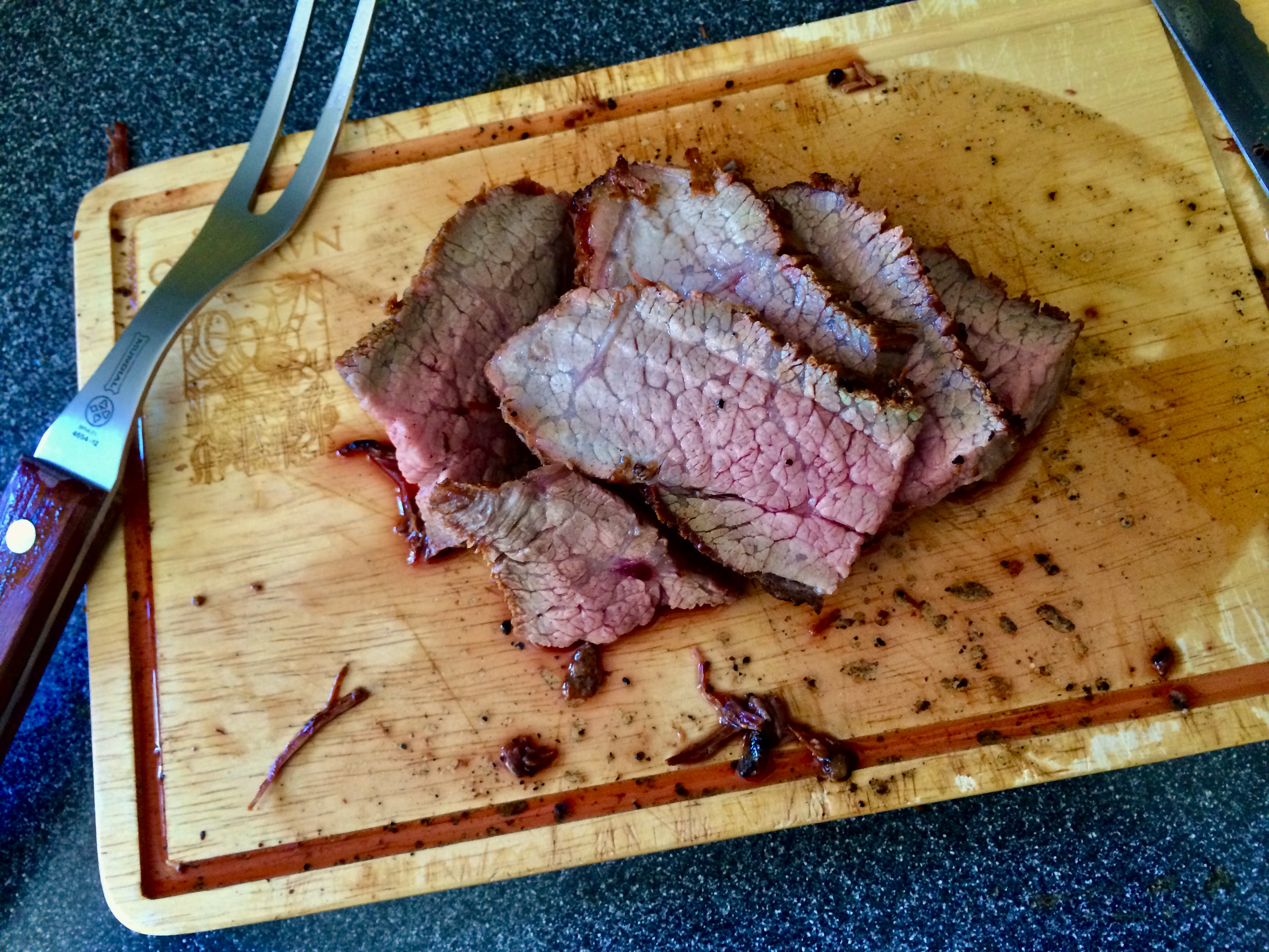 Sliceable slow-cooked tri-tip resting on a cutting board with visible grain and a small bowl of strained onion-garlic broth nearby