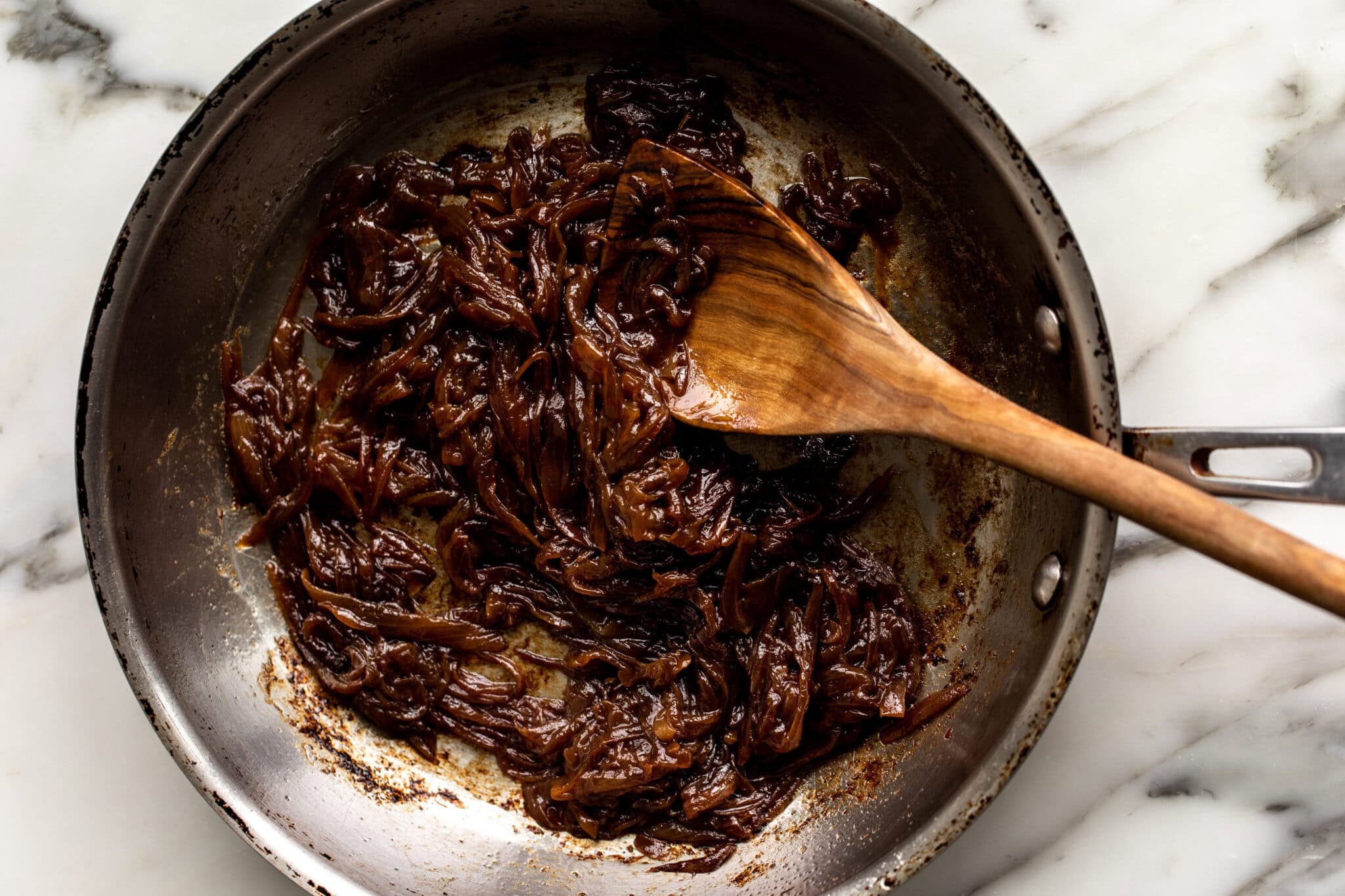 Thinly sliced onions cooking in a stainless steel skillet while a spoon scrapes browned bits from the bottom