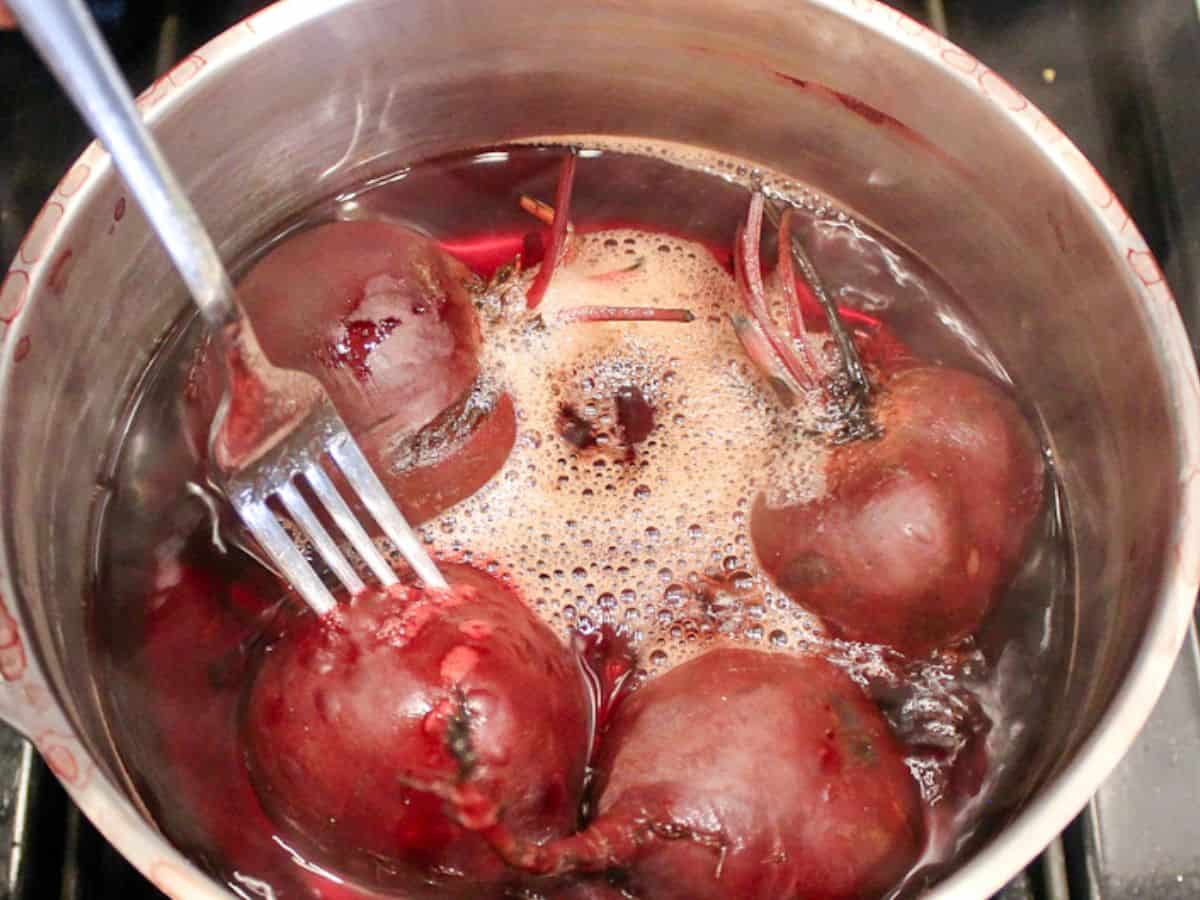 Whole beets simmering in a stainless steel pot on a stovetop with gentle bubbles and steam rising